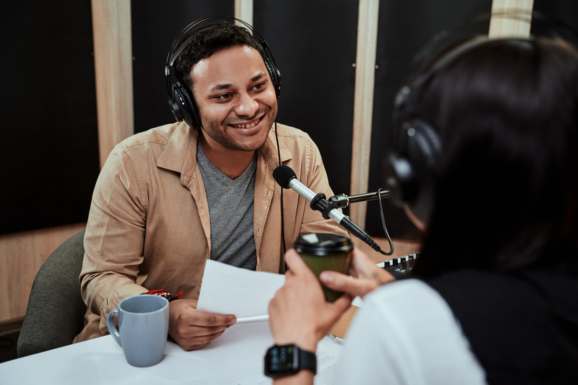 Portrait of young male radio host going live on air, talking with female guest, holding a script paper while sitting in studio